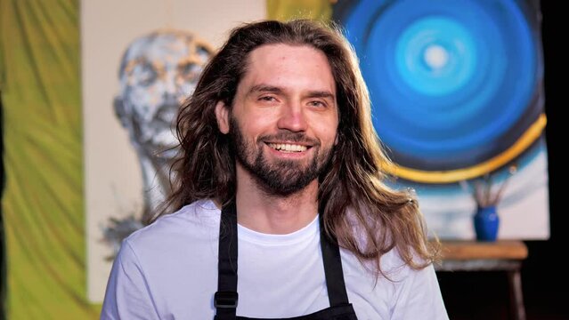 Professional Young Male Artist with Long Hair, Healthy White Teeth, Charming Smile, Wearing Apron, Looks at the Camera with a Smile. Authentic Creative Studio with Large Canvas. Face Portrait. 