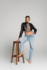 Fototapeta premium full length of fashionable african american model in cropped jacket posing with hand on hip near high chair on grey.