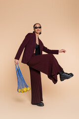 full length of stylish african american woman in burgundy suit and sunglasses holding mesh bag with lemons on beige.