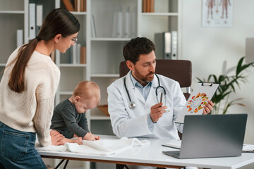 Fototapeta premium Having consultation. With baby and mother. Doctor in formal clothes is working in the cabinet