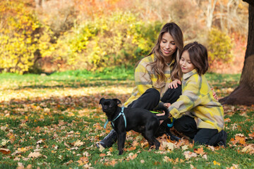 Mom and daughter with doggy in yellow coats spend time together in autumn park outdoors, having fun. Cute kid with mother posing in fall nature. Concept of family leisure activity. Copy ad text space