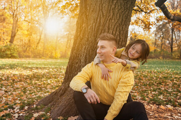 Dad and daughter in yellow clothes spend time together in autumn park outdoors, happy hugging having fun. Adorable kid with father near tree in fall nature. Concept of family leisure. Copy text space