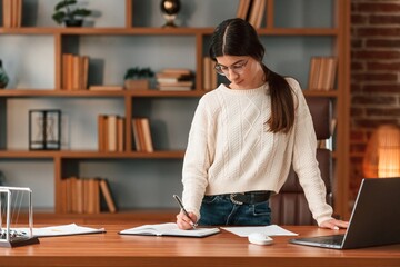 Young woman in glasses is working indoors. Standing by table with laptop