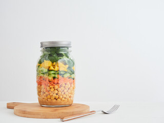 Glass jar with multi-layered salad of chickpeas and vegetables for a healthy lunch. Cucumber, carrot, mango and lambs lettuce. Healthy food, diet, detox, clean eating and vegetarian concept.