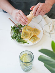 Partial view of adult woman at table with wooden pepper grinder in her hands, grinding pepper over plate of pasta (ravioli) and arugula salad.