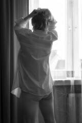 Woman at the window in a pink shirt. A woman looking out the window in a men's shirt and briefs. Black and white version - obrazy, fototapety, plakaty
