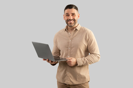 Happy middle aged businessman in shirt chatting with client by laptop , smiling at camera on grey background, free space
