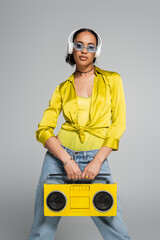 stylish african american woman listening music in wireless headphones and holding retro boombox isolated on grey.