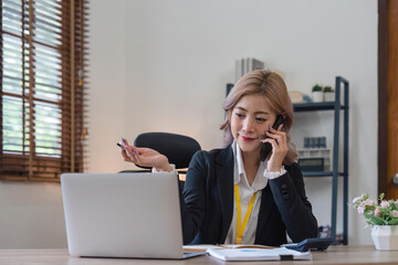 Happy young asian woman talking on the mobile phone and working smiling while sitting at her working place in office