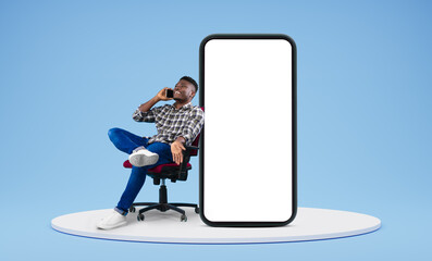 Laughing young african american guy call by smartphone, talk, sit in armchair with huge smartphone