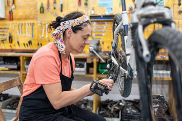 woman repairing bicycle hanging in bicycle workshop wearing black apron, salmon T-shirt and headscarf