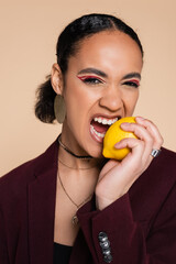 african american woman in burgundy blazer biting ripe lemon isolated on beige.