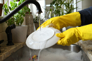 Washing rinsing dishes in kitchen sink by hand wearing yellow rubber gloves.