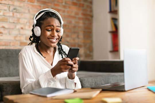 Portrait of smiling black woman sitting at desk with laptop, using cellphone and wearing headphones, free space