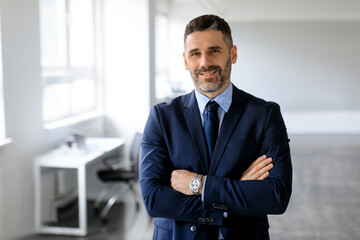 Portrait of confident middle aged male entrepreneur standing with folded arms and smiling at...