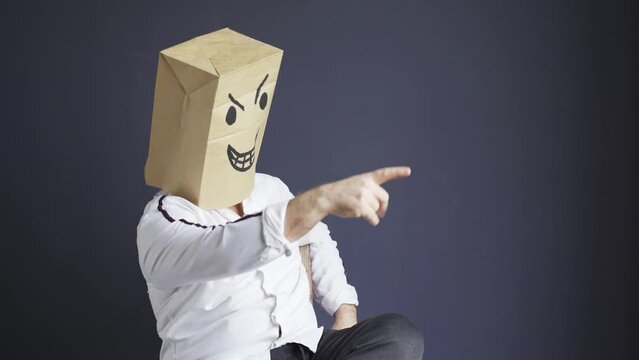 A man in a white shirt with a bag on his head, with a drawn angry emoticon, emotionally gloats while sitting on a chair. Laughing, gesturing with his hands. Emotions and gestures.