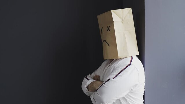 A sad man in a white shirt with a bag on his head, with a sad emoticon drawn, stands against the wall, cries, turns around. Emotions and gestures.