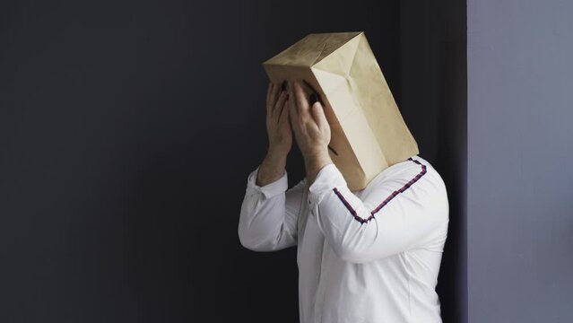 A sad man in a white shirt with a bag on his head, with a drawn crying emoticon, stands near the wall, holds his head and cries. Emotions and gestures.
