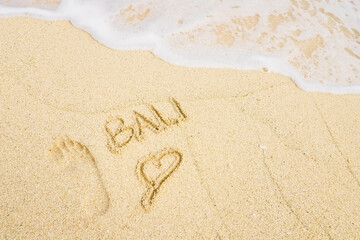 The word BALI is writting on the sand with love sign, footprints, and wave. drawing on the sand. writing with sands on the beach. 