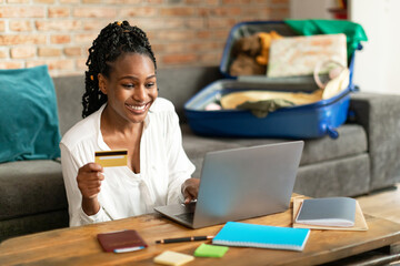 Happy black lady booking vacation at travel agency or making hotel reservation online, using laptop and credit card