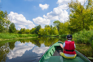 A young boy in a baseball cap and a red life jacket on a green boat is sailing among the trees on a sunny day. Summer. - 586641467