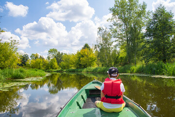 A young boy in a baseball cap and a red life jacket on a green boat is sailing among the trees on a sunny day. Summer. - 586641613