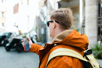 Man wearing in travel orange jacket, standing on roadside in city holding smart phone in his hand...