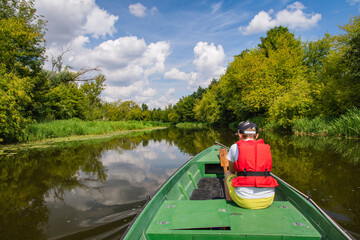 A young boy in a baseball cap and a red life jacket on a green boat is sailing among the trees on a sunny day. Summer. - 586643814