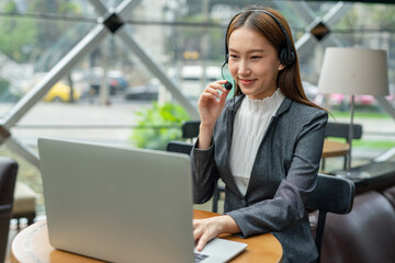 Young asian businesswoman working on her computer laptop and headset microphone in a cafe. Customer service executive working out of home. Hybrid workplace.