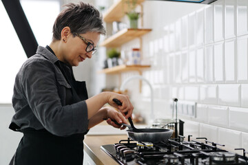 An older woman in a black apron is frying dinner in a frying pan in the home kitchen.