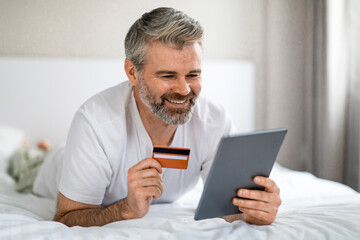 Positive mature man enjoying online shopping, lying on bed