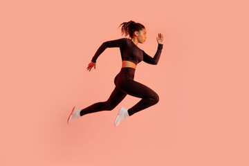 Fototapeta na wymiar Sporty black lady jumping or running, posing in mid-air, exercising over peach neon background, side view, full length