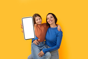 Smiling Young Mother And Little Daughter Demonstrating Smartphone With Blank White Screen