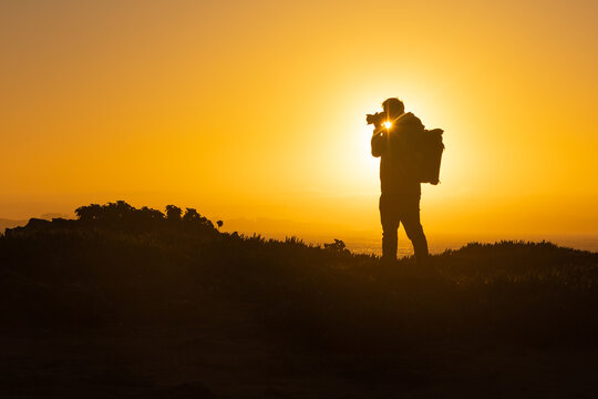 A silhouette of a man standing on top of a cliff and taking photos of sunset