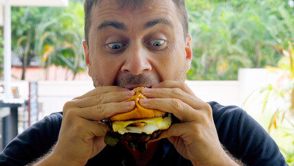 Man eat juicy hamburger at an outdoor restaurant. Man enjoys delicious and unhealthy meal causes funny and emotional expressions, happily holds the big burger with two hands and takes big bite