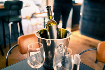 Wine bottle in ice bucket on restaurant or bar table. Drink cooler in pub. White sauvignon blanc, chardonnay or riesling. Party, anniversary celebration or date. Waiter or sommelier in the background.