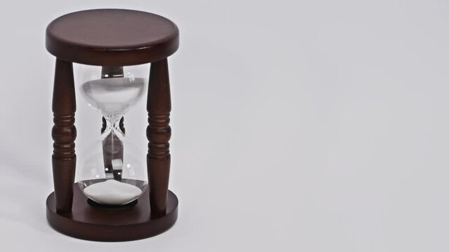 White sand trickle in hourglass, sand watch isolated on white background, wooden dark brown three legged stand, time is passing, antique device for measuring time. Close up, 4K Ultra HD.