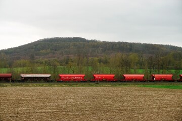 Fototapeta na wymiar Grünes Landschaft Panorama mit braunem Ackerfeld vor Zug mit roten Güterwaggons vor Wald und Himmel am Mittag im Frühling