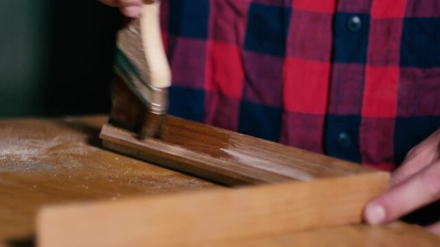 Handyman worker in a red plaid shirt paints a wooden frame with varnish with a brush in a carpentry workshop.