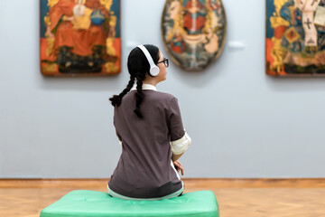 Back view of young Caucasian pretty woman wearing headphones and contemplates ancient artefacts. Student visiting gallery or museum. Concept of education and culture