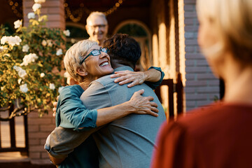 Happy senior woman embraces her son while welcoming him and his wife in front of house.