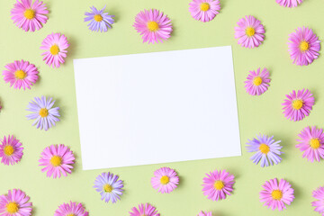 Greeting card template. Pink and purple flower aster on green table background with blank paper sheet. Flat lay, top view, mockup, copy space for text. Aesthetic stylish floral pattern.