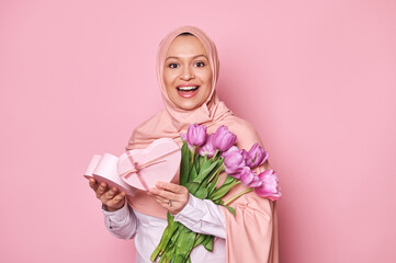 Cheerful Arab lady in pink hijab, holding a heart shaped gift box and tulips, smiling to camera. Positive amazed Muslim woman with Mother's Day or birthday present posing over pink studio background