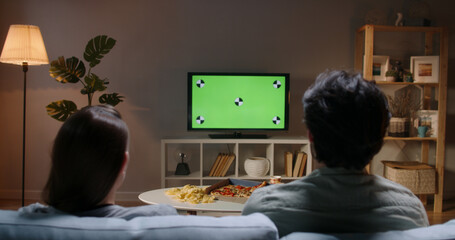 Couple spending time together in the evening, sitting on couch and watching tv with green...