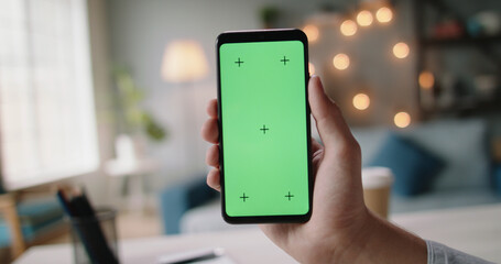 Hand of guys holding a smartphone with green chroma key screen in living room. Template for online store or social media 