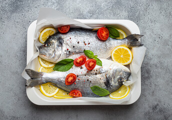 Two raw fishes dorado in casserole dish with ingredients lemon, fresh basil, cherry tomatoes close up top view on rustic stone grey background, cooking healthy fish dorado concept
