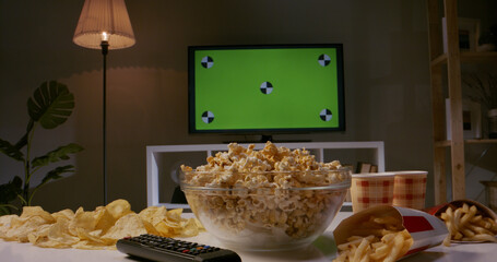 Closeup shot of tv set with green chromakey screen on wall and lots of popcorn, french fries and chips on table ready for watching favourite tv shows 