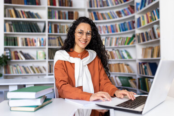 Portrait of cheerful smiling girl student, woman looking at camera sitting studying with laptop, Hispanic woman wearing glasses studying online in university library among glasses using laptop.