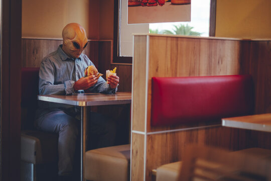 One man with alien mask eating alone inside a fast food store bad food hamburger and french fries. Ufo are among us living as human concept funny image. Invasion and diversity people indoor leisure