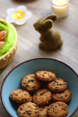 Easter eggs in the basket, bowl of cookies, chocolate pralines, Easter bunny figurine, cups of tea, glasses of juice, flowers and lit candles on the table. Selective focus.